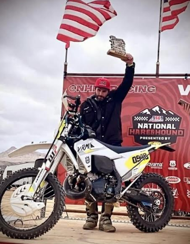 Motocross rider on podium raising a trophy, with flags and race banner in the background.