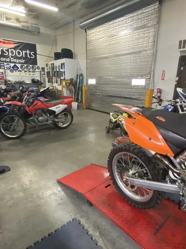 Motorcycles in a workshop, including red and orange dirt bikes on a concrete floor and red mat.
