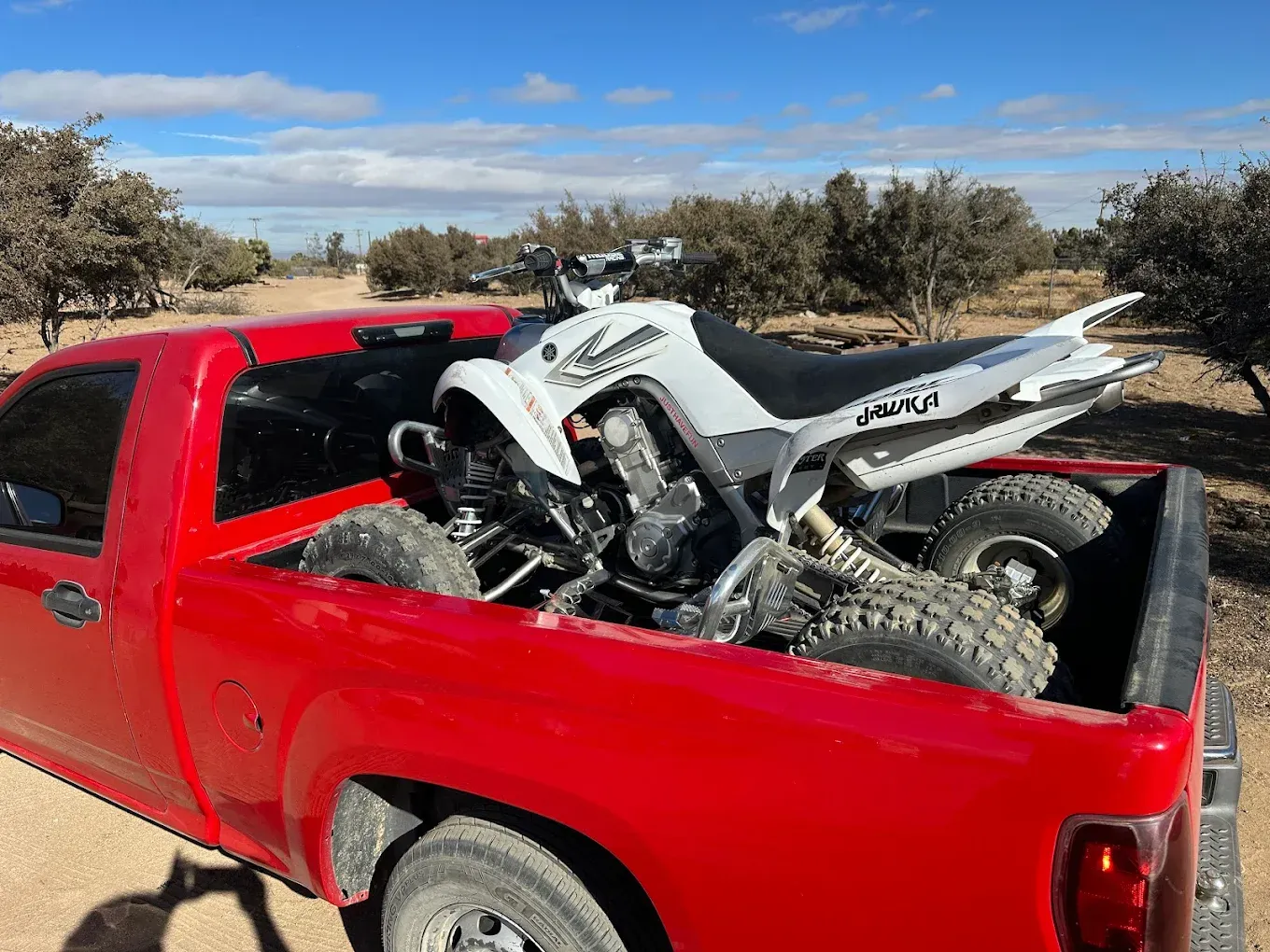 Red pickup truck hauling a white dirt bike in its bed on a dusty road.