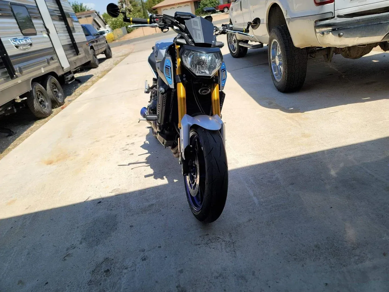 Blue-and-black sport motorcycle parked on a sunlit driveway between pickup trucks