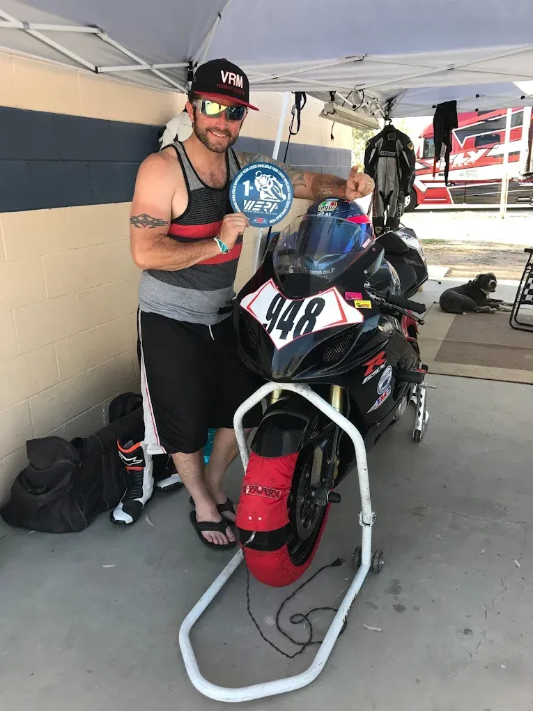 Man standing beside a red and black motorcycle under a tent, holding a blue sign