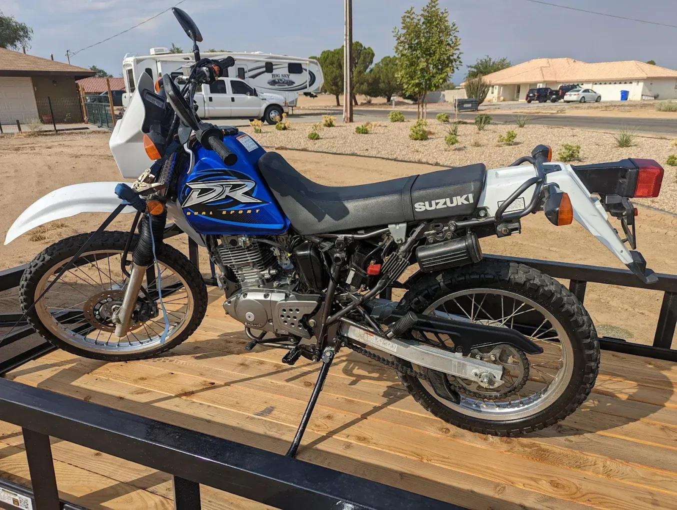Blue and white dual-sport motorcycle secured on a wooden trailer in a sunny outdoor lot