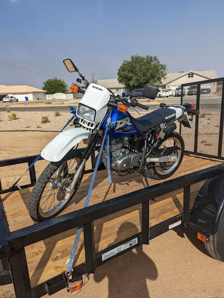 Blue dirt bike strapped on a trailer in a sandy outdoor lot under a clear sky.