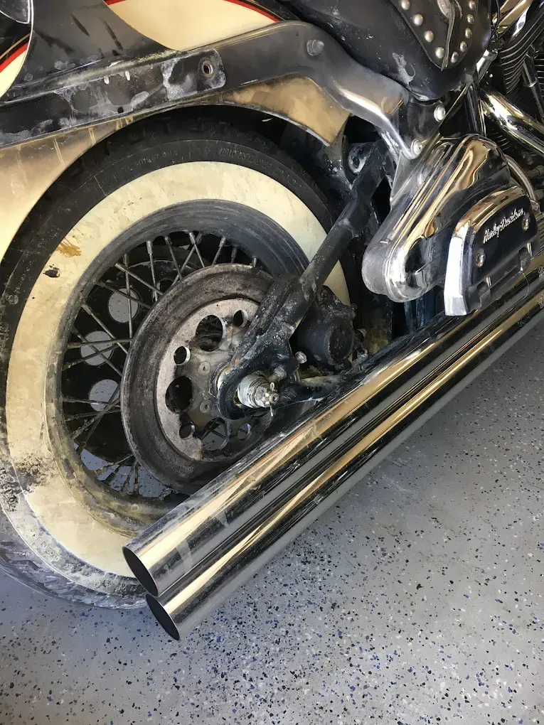 Close-up of a motorcycle’s rear wheel, engine, and chrome exhaust with whitewall tire on a garage floor.