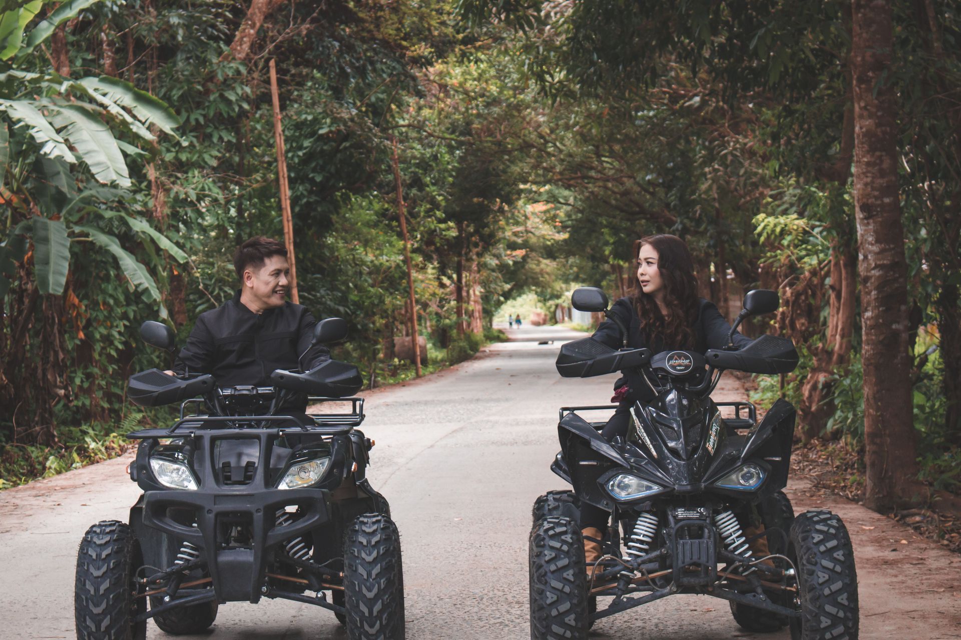 Two people on ATVs facing each other on a forest road, surrounded by trees.