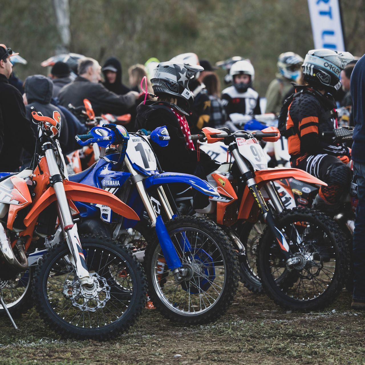 Motocross riders lined up on dirt bikes at a starting gate, with orange and blue bikes in the foreground