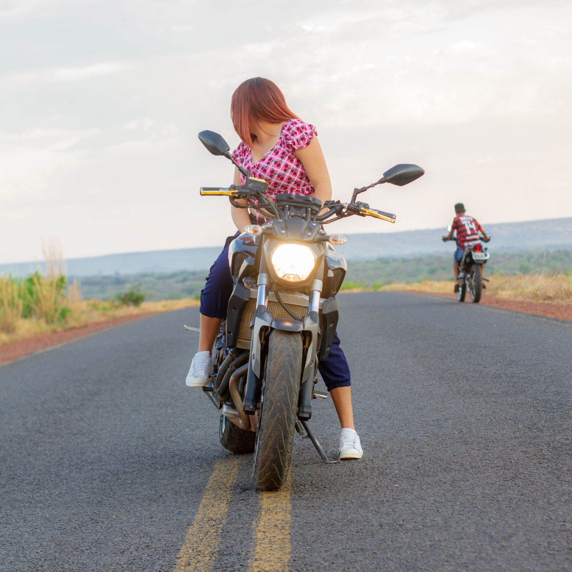 Two motorcyclists on a rural road, with a rider in a pink shirt looking down at the bike.