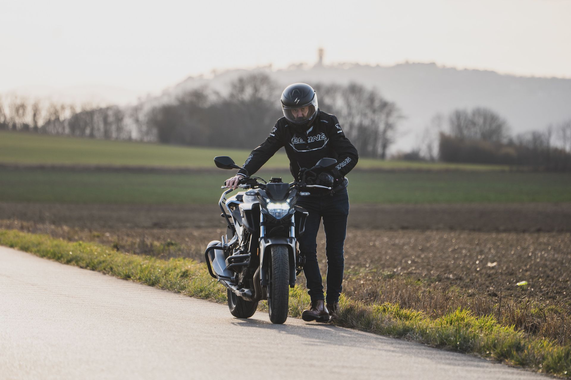 Motorcyclist in black gear pushing a bike beside a rural road at dusk