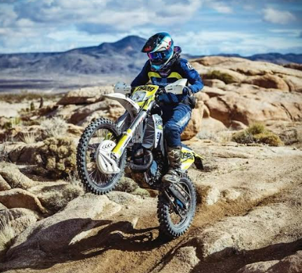 Dirt biker in blue gear riding over rocky desert terrain with mountains in the background