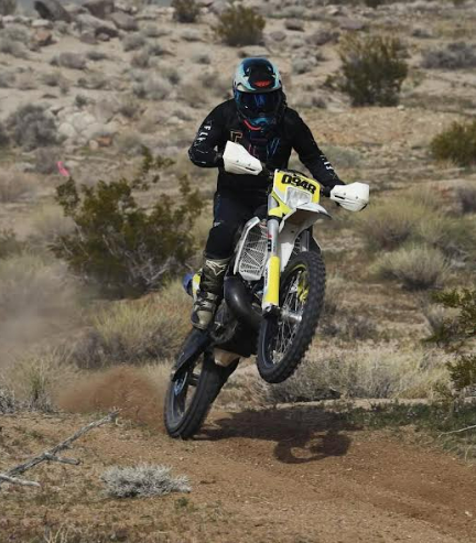 Motorcyclist in black gear rides a yellow dirt bike through a rocky desert trail, front wheel lifted.
