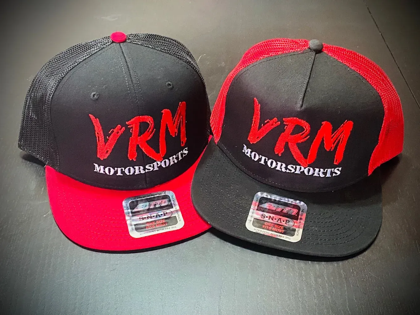 Two black-and-red VRM Motorsports baseball caps side by side on a dark surface