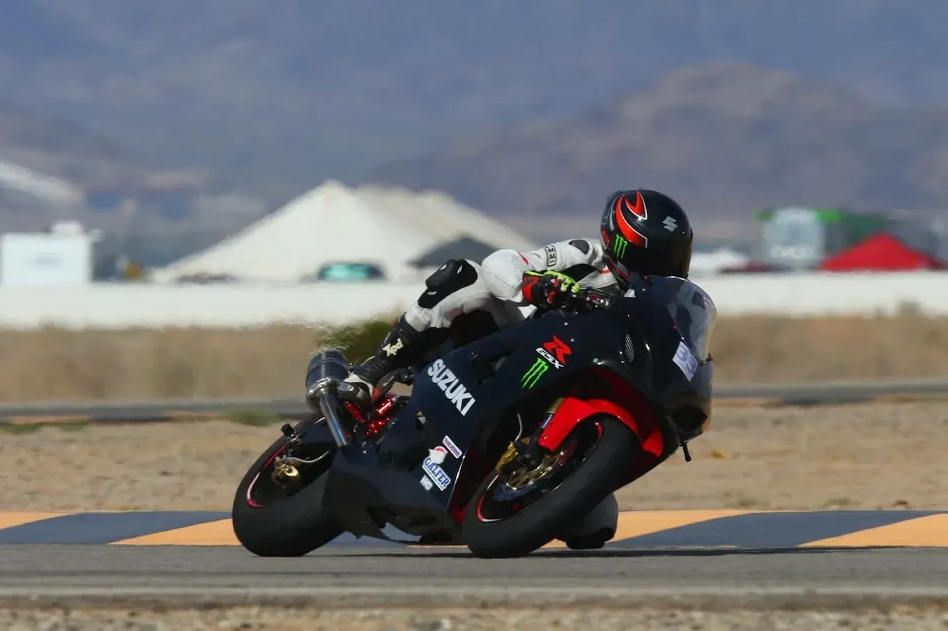 Motorcyclist leaning hard into a turn on a racetrack, black bike with red accents, desert mountains behind.