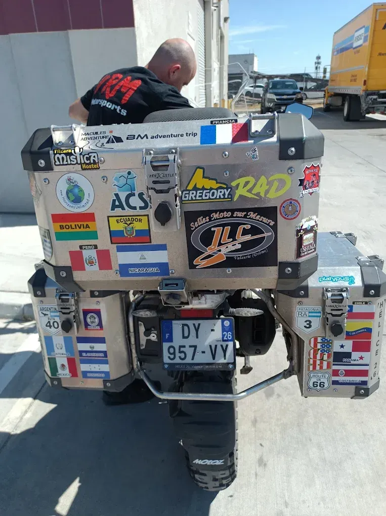 Sticker-covered motorcycle parked on a sidewalk beside a road, with a person leaning over it.